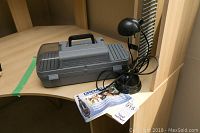 Grey plastic Dremel carrying case and black desk lamp on light wood desk.