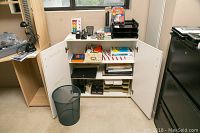 Wide view of office cabinet with white doors open showing various office supplies on shelves and top surface, including trays, paper packages, and a black mesh wastebasket in front.
