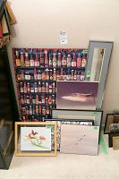 Seven framed art pieces leaned against a wall including colorful hot sauce bottles display, jet aircraft photo, watercolour painting, ceramic tile with cracks and other art pieces.