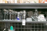 Shelf with clear glassware including wine glasses, champagne glasses, flute glasses, glass decanter, shot glasses, and small bowls.