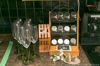 Photo of 9 champagne glasses with green stems lined up on countertop near wooden knife block and spice rack with glass jars. Two pairs of decorative salt and pepper shakers are visible.