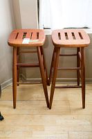 Pair of two wooden stools with slatted rectangular seats and turned legs, shown standing on a wood floor near a window.