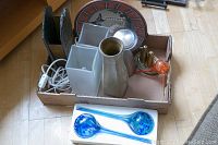 Overview of all items in the lot arranged in a cardboard box on a wooden floor showing two white frosted glass vases, tall grey ceramic vase, decorative plate at the back, metal salt and pepper shakers, and colored glass water bulbs in front.