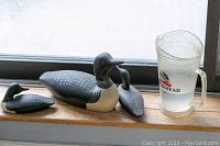 Three decorative loon figurines placed on a windowsill next to a plastic Measuring pitcher; loons show black and white painted details; one smaller loon with neck damage appears repaired