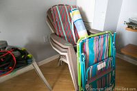 Pair of folding lawn chairs stacked with three brown resin Muskoka chairs stacked against wall with cushions and neck roll