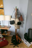 View of six purses and two knap sacks hanging on a coat rack in a corner of the room. Includes table with lamp and other bags on floor.
