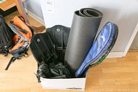 Box with scuba fins, exercise mat, and rackets in blue case on wooden floor near backpack