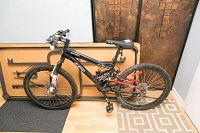 Side view of black CCM Static 24 mountain bike leaning against a folded table, showing full suspension with red rear suspension components and knobby tires.