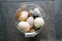 Six decorative stone eggs and one amethyst cluster arranged inside a clear glass pedestal container, viewed from above on tile floor.