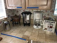 Wide view showing all items arranged on a kitchen counter: stainless steel thermos, vintage copper pot with warmer, coffee urn, Gevalia coffee maker, and boxed Cuisinart food processor.