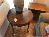 Wood round side table with wood grain finish and curved legs positioned between two upholstered chairs, with a large decorative lamp on top.