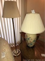 Floor lamp standing beside beige curtain next to matching ceramic table lamp on round wooden table, both with cream pleated shades.