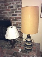 Photo showing both lamps set on a brick hearth with visible power cords. The taller lamp features beige cylindrical shade and horizontally striped ceramic base in dark and light tones. The shorter lamp has a white pleated shade and rounded dark brown ceramic base with decorative patterning.