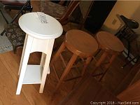 Three wooden stools shown in a room with hardwood floor. Two natural wood round stools and one white painted stool with open base.