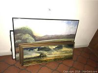 Four framed landscape paintings stacked against wall, showing variety in size and frame style.