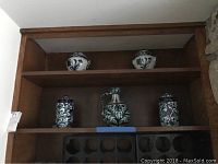 Five blue and white pottery pieces displayed on wooden shelves, including two small pots on top shelf, large handled pitcher and two jars with lids on middle shelf.