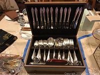 Wooden flatware box with knives in the lid slots and various spoons, forks, and serving utensils in the base.