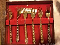 Close-up photo of one set of 6 gold-plated serving forks in a wooden box with red velvet lining, showing ornate, detailed handles and 4 tines per fork.
