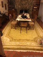 Full view of the large hand-woven area rug placed under a dining table with 6 chairs and visible stone fireplace background.