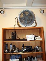 Wooden shelf with multiple cordless phones, base units, chargers, cables, and electronic accessories. Large 18-inch fan sits on top shelf against wall.
