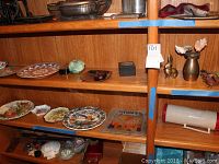 Three wooden shelves filled with various decorative plates, a green silk flower, small boxes, a brass-style duck figurine, and a brass-style vase with artificial flowers.