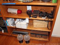 Wooden shelving unit with various personal electronic devices, chargers, cassette storage boxes, a woven basket, and two small lamps on lower shelf.