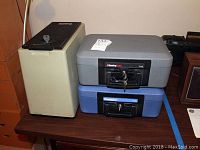 Three lock boxes on a wooden surface: two SentrySafe lock boxes (gray 1110 and blue 1100) and a beige box with a black lid and lock mechanism.
