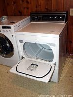 White Kenmore electric dryer with front drop-down loading door open, showing interior drum next to another washing machine, placed against wood-paneled wall.