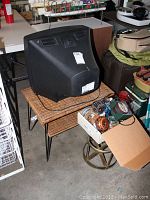 Wicker table with black CRT television on top and box of electronics and decor items below on floor.