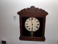 Front view of the vintage wooden wall clock mounted on the wall, showing clock face, pendulum through glass door, and carved wooden frame.